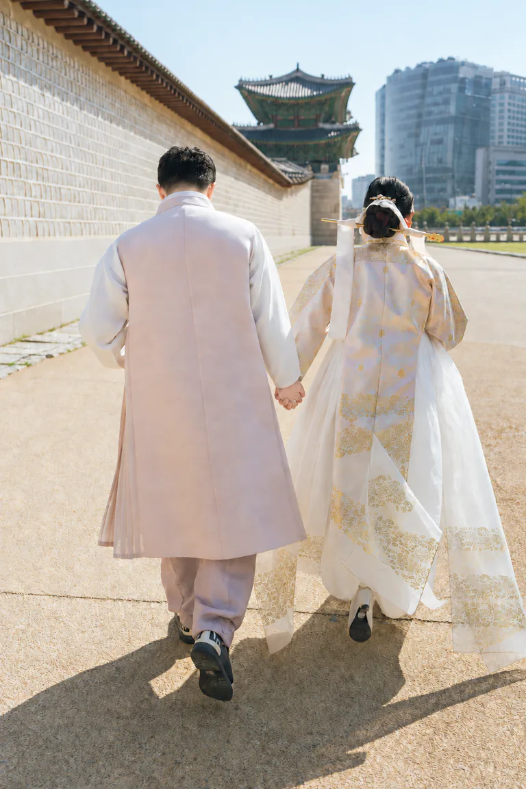 한복을 입은 두 사람이 손을 잡고 서울의 궁궐 담장 옆을 걷는 뒷모습